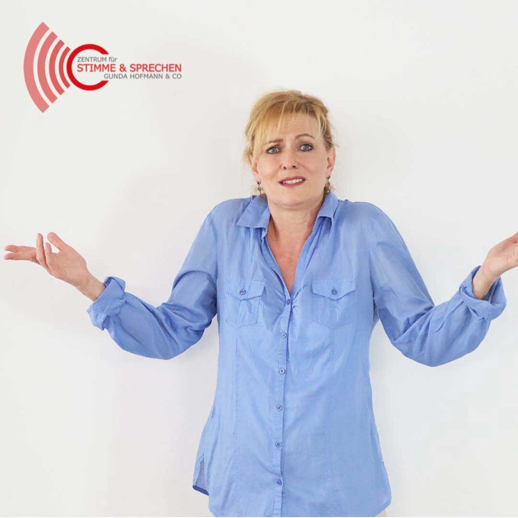verstanden-werden-Stimme und Sprechen Gunda Hofmann - Zentrum für Stimme & Sprechen Blonde woman in a blue button-down shirt shrugs her shoulders, with a red logo top-left reading 'Zentrum für Stimme & Sprechen' on a white wall.