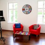 seminarraum-mieten-raum1-beratung-25 - Zentrum für Stimme & Sprechen Bright room with two red armchairs, a colorful striped coffee table, a round multicolored clock, and a photography light on a stand.