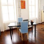 seminarraum-mieten-raum1-beratung-26 - Zentrum für Stimme & Sprechen Bright home studio with a black table, two blue chairs, a laptop, and a music stand with a light on a tripod by large windows.