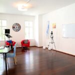 seminarraum-mieten-raum1-kamera-27 - Zentrum für Stimme & Sprechen Bright studio room with a black desk, blue chairs, red armchairs, a camera tripod, and a whiteboard on the wall by two windows.