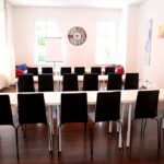 seminarraum-mieten-raum1-klassenzimmer-5 - Zentrum für Stimme & Sprechen Bright learning room with white tables in rows and black chairs, a wall clock centered between windows at the back.