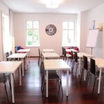 seminarraum-mieten-raum1-klassenzimmer-7 - Zentrum für Stimme & Sprechen Bright classroom with white tables and black chairs arranged in rows, a wooden floor, two windows, a colorful wall clock, whiteboard, flip chart, and a red seating area at the back.