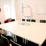 seminarraum-mieten-raum1-meeting-raum-1 - Zentrum für Stimme & Sprechen Bright conference room with a large white table surrounded by black-and-wood chairs, a colorful wall clock, and a whiteboard on the wall with windows letting in natural light.