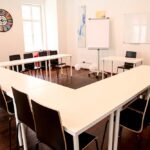 seminarraum-mieten-raum1-meeting-raum-3 - Zentrum für Stimme & Sprechen U-shaped meeting room with white tables and black chairs, bright with natural light, whiteboard at front and flip chart nearby.