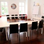 seminarraum-mieten-raum1-meeting-raum-4 - Zentrum für Stimme & Sprechen U-shaped conference room setup with white tables, black chairs, and wooden floor, bathed in natural light from two windows.