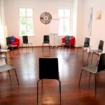 seminarraum-mieten-raum1-sesselkreis-8 - Zentrum für Stimme & Sprechen Circle of chairs arranged in a bright room for group discussion or meeting, with two red chairs and a flip chart at the back.
