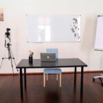 seminarraum-mieten-raum1-video-23 - Zentrum für Stimme & Sprechen Camera on a tripod aimed at a desk with a laptop, in a bright studio featuring a whiteboard and a rolling flip chart nearby.
