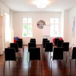 seminarraum-mieten-raum1-vortrag-13 - Zentrum für Stimme & Sprechen Bright meeting room with rows of black chairs facing a flip chart and a colorful circular clock on the back wall.