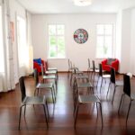 seminarraum-mieten-raum1-vortrag-21 - Zentrum für Stimme & Sprechen Bright, sunlit room set up for a talk with two rows of gray chairs on a wooden floor, red lounge chairs in the back corners, and a colorful wheel on the wall center.