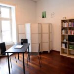 seminarraum-mieten-raum2-15 - Zentrum für Stimme & Sprechen Small study room with a black table and two chairs near a large window, next to a tall white room divider and a bookshelf.