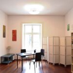 seminarraum-mieten-raum2-17 - Zentrum für Stimme & Sprechen Bright minimalist study room with a window, a black table and two chairs, a lamp, and folding room dividers against white walls.