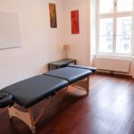 seminarraum-mieten-raum2-behandlungsliege-9 - Zentrum für Stimme & Sprechen A massage table in a bright, minimal therapy room with a wooden floor, a whiteboard on the wall, and a window letting in natural light.